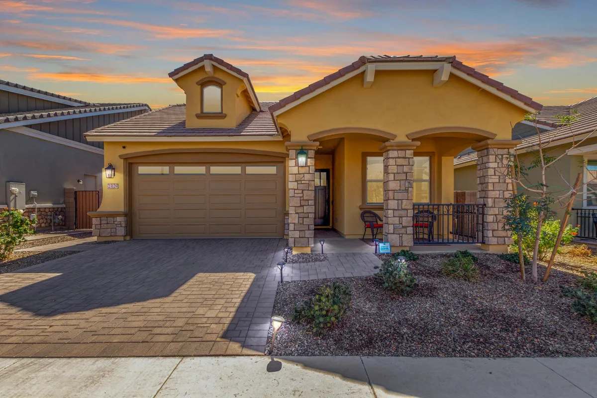 The image showcases the inviting exterior of a single-family home in Queen Creek, Arizona, featuring a charming front porch framed by elegant stone columns, a spacious garage, and lush, manicured landscaping. The warm Arizona sunset bathes the scene in a golden glow, hinting at relaxing evenings spent outdoors.