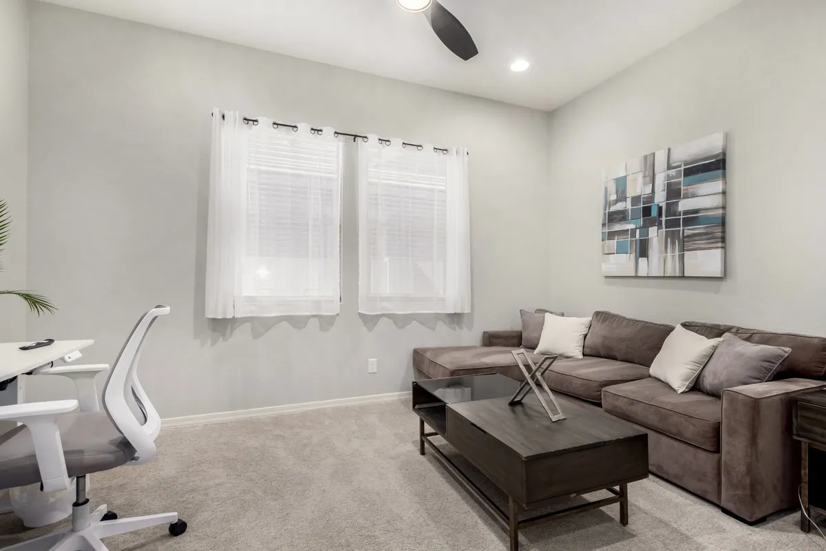 This image shows a tranquil and productive den/office space featuring a desk with chair, a comfortable sofa, a coffee table, and artwork on the wall. Natural light fills the room, enhancing the atmosphere for focused work or quiet reflection. The layout offers a dedicated workspace while maintaining a cozy and welcoming feel.