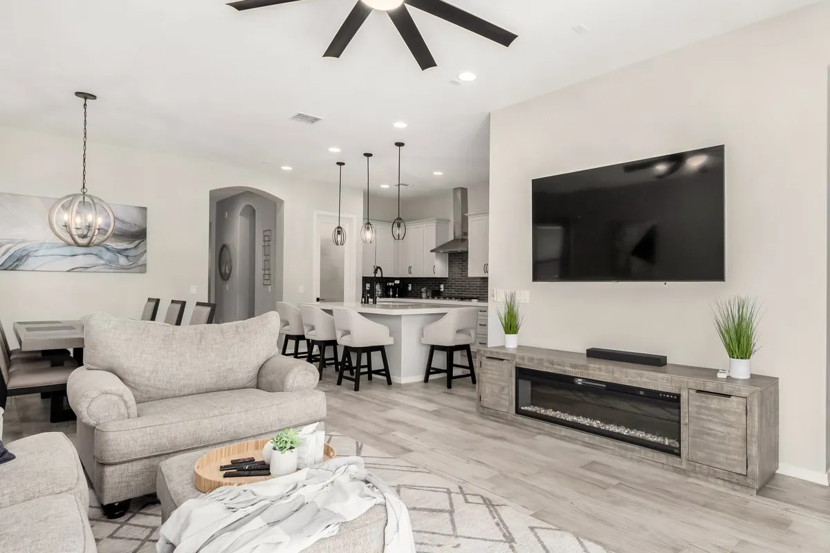 This image captures a welcoming open-concept layout with a comfortable gray sofa, a modern dining area with an elegant light fixture, a large flat-screen TV, and a kitchen island with bar stools. The connected living, dining, and kitchen spaces are bathed in natural light, emphasizing the seamless flow between each area.