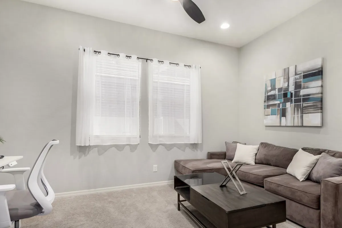 The scene features a contemporary and serene room with a sofa, coffee table, windows that allow sunlight to pour in, and wall art. The setup invites both relaxation and work-from-home activities, making it ideal for flexible use as a den or office. The emphasis on natural light and comfortable furnishings aligns with modern home office aesthetics.