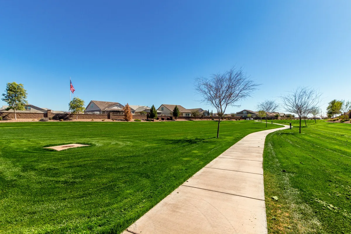 A tranquil neighborhood park scene featuring a manicured lawn, mature trees, and a broad sidewalk perfect for walking or jogging. Homes are visible in the background, flanked by a waving flag, conveying a sense of community and open space.