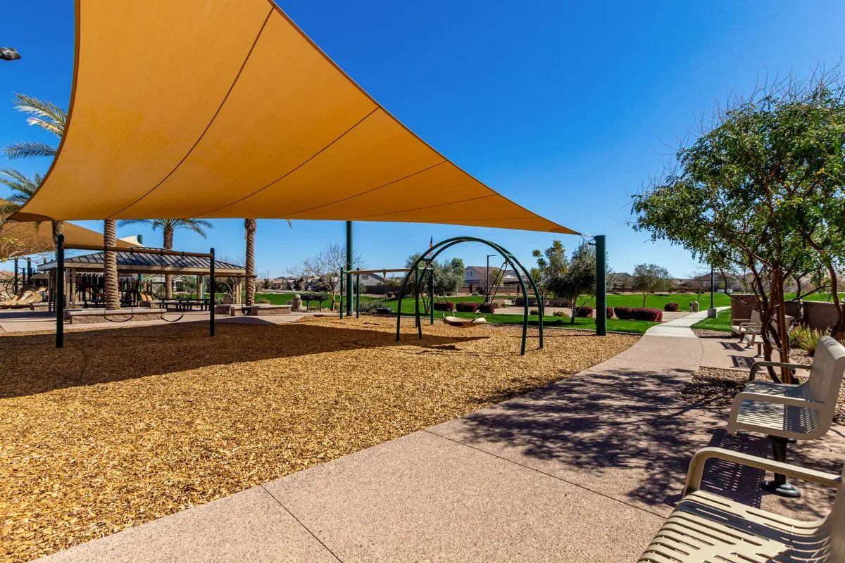 This image showcases a vibrant community park area with a green lawn, a shade structure, swings, and families or children playing. The scene captures a lively atmosphere with people walking, socializing, and enjoying outdoor activities in a communal space.