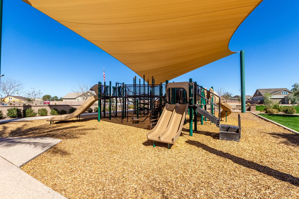 A vibrant playground structure equipped with slides sits under a protective shade sail, surrounded by wood chips and open space. Children are visible playing and laughing, with a few families gathered nearby, suggesting a lively community park atmosphere.