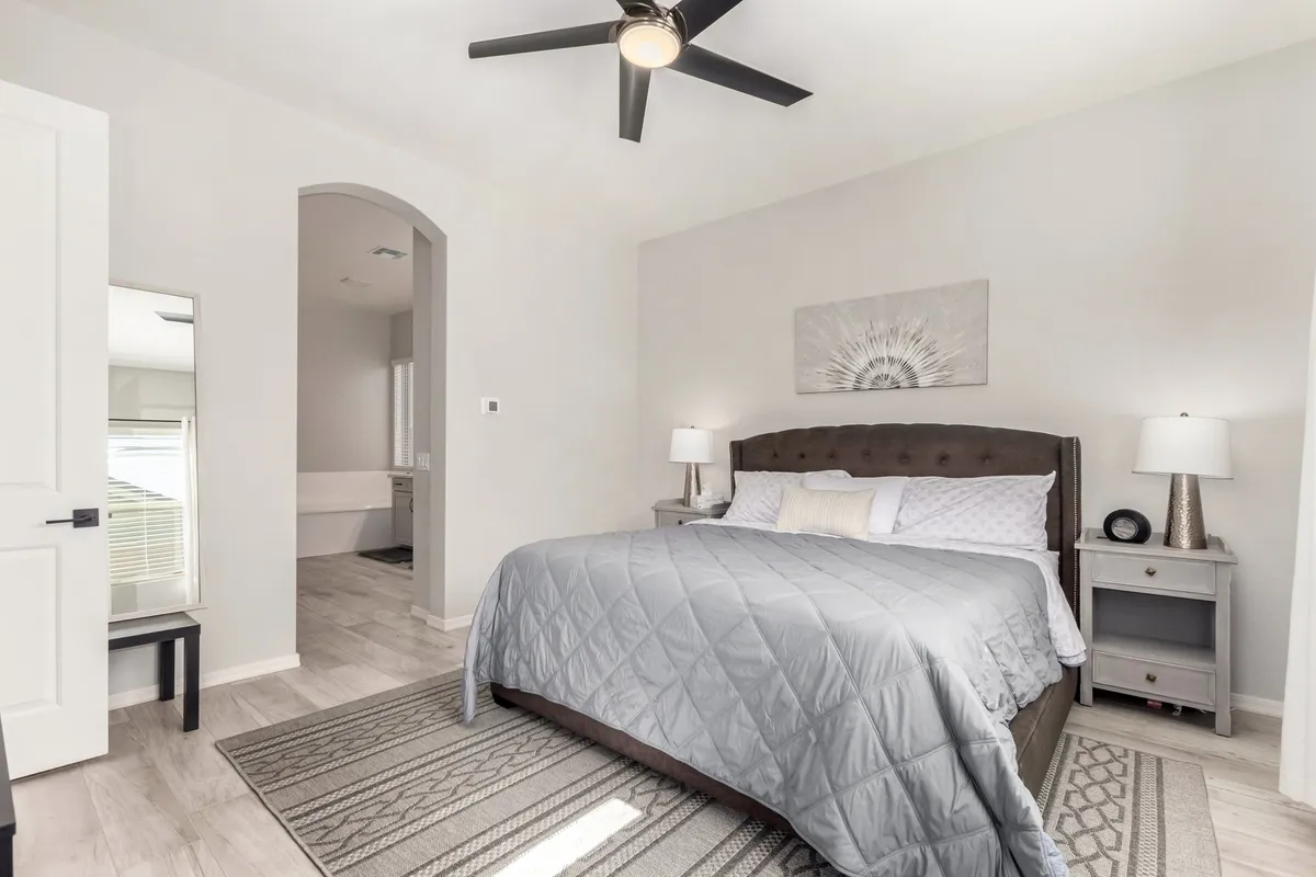 A spacious bedroom featuring a neatly made bed with coordinating nightstands, soft ambient lighting, a large mirror, and tasteful artwork adorning the walls. The overall atmosphere is calm and inviting, ideal for rest and relaxation.