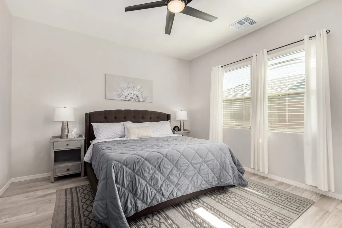 A tranquil bedroom with a bed dressed in a gray quilt, a nightstand with a modern lamp, sheer curtains framing generously-sized windows, and decorative wall art. The space exudes a peaceful, restful vibe with plenty of natural light.